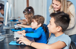 Children are sitting  at a desk with computers and a teacher is looking at them 