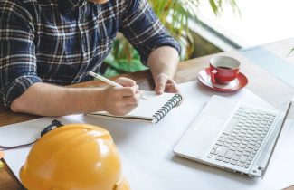 Henkilö kirjoittaa kannettavan tietokoneen äärellä lehtiöön vierellään pöydälle laskettu turvakypärä.