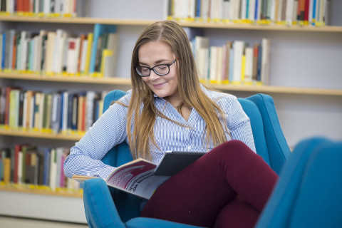 Nuori nainen istuu nojatuolissa ja lukee oppikirjaa.