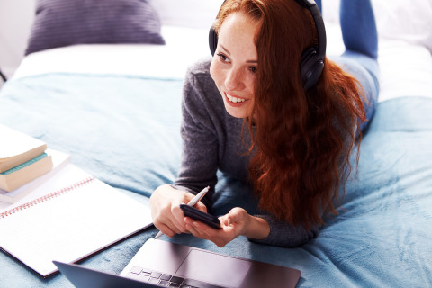 Young woman on a bed, laptop, notebook and notes in front of her, holding a mobile phone.