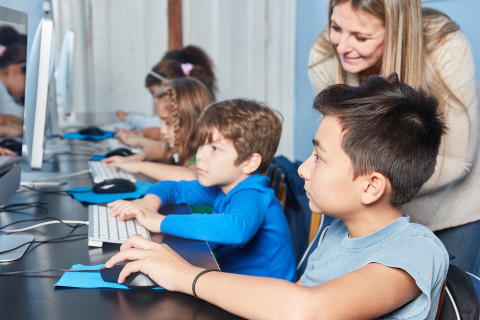 Children are sitting  at a desk with computers and a teacher is looking at them 