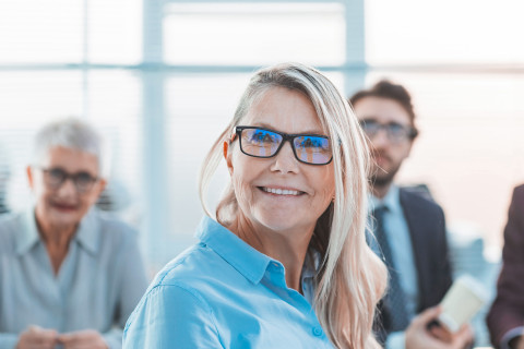 A lady is smiling and staring at somewhere. There are people sitting behind her.