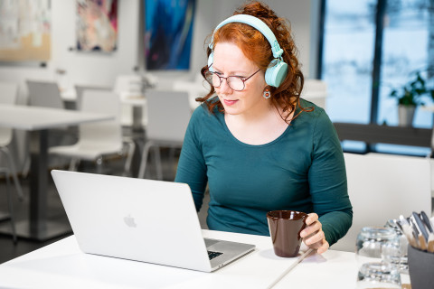 nainen opiskelee tietkoneen edessä kuulokkeet korvilla ja kahvikuppi kädessä