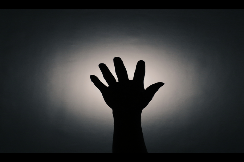 Mustalla taustalla keskellä valoisampi kohta jonka edessä ylöspäin kurottava käsi sormet harallaan.