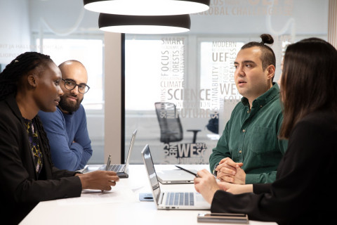 Neljä henkilöä neuvottelee pöydän äärellä aulatilassa.