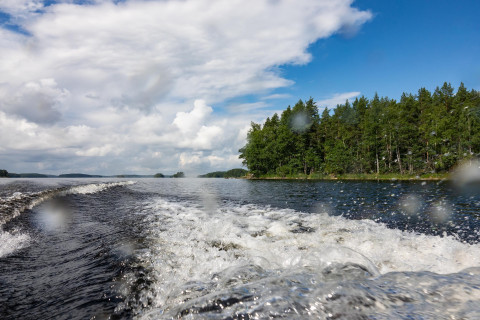 Veneen perävana järven pinnassa. Taustalla saari ja pilvinen taivas.