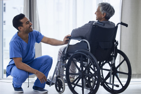 Siniasuinen hoitaja kyykyssä pyörätuoliasiakkaan edessä
