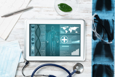 Tabletti näyttää ihmisen siluetin ja terveystiedon graafeja, ympärillä stetoskooppi, maski ja röntgenkuvia.