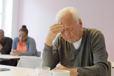 Ikääntynyt henkilö nojaa otsaa käteensä luokkahuoneen päydän ääressä.