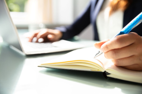 Henkilö kirjoittaa muistivihkoon taustalla kannettava tietokone, jonka näppäimistöllä toinen käsi on.