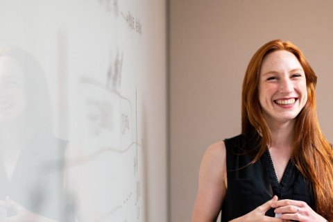 Punahiuksinen hymyilevä nainen seisoo valkotaulun edessä