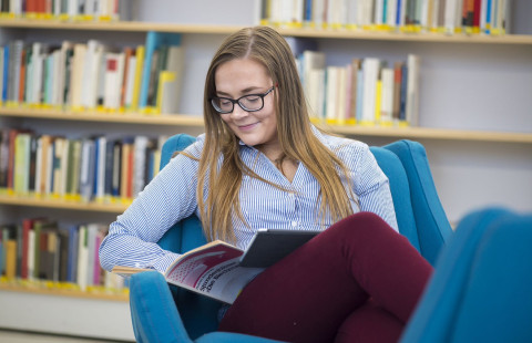 Nuori nainen istuu nojatuolissa ja lukee oppikirjaa.