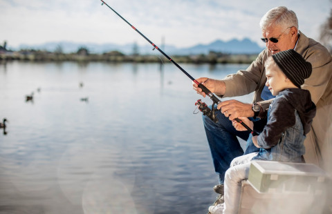 An old man and a young boy sitting on a pier and trying to catch fish.