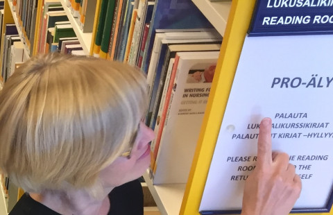 Kirjaston työtekijä osoittaa hyllynpäätykylttiä, jossa lukee aakkosvälitieto pro-äly.