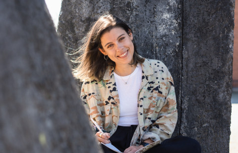 Young woman sitting outside, pen and notebook on her lap.