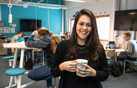 Young woman looking at the camera smiling and holding a coffee cup, young men working with their laptops in the background.