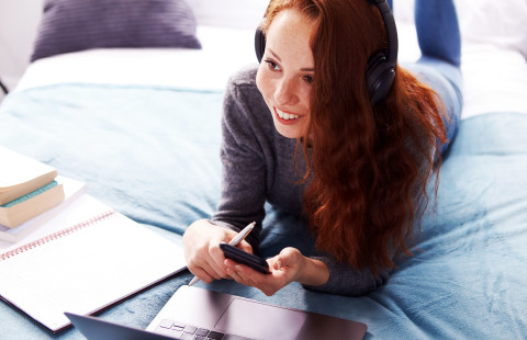 Young woman on a bed, laptop, notebook and notes in front of her, holding a mobile phone.