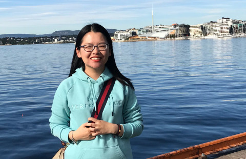 Woman with dark hair and glasses smiling for the camera, the photo is taken by the sea or other water.