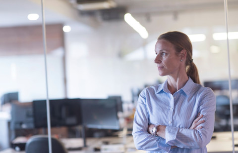 Nainen seisoo toimistossa ja katsoo sivuun kamerasta