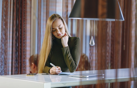 Nainen istuu korkean pöydän äärellä ja kirjoittaa muistiinpanoja vihkoon