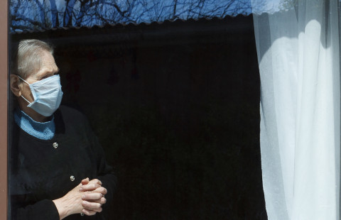 Old woman wearing a mask looking through a window.