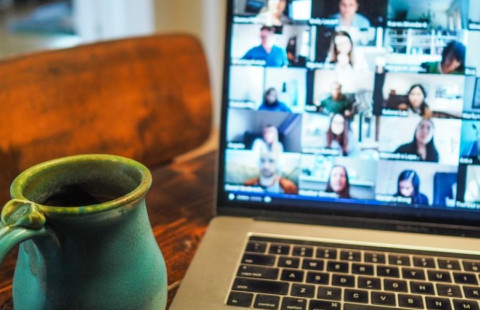 Tietokone, näytöllä kuva verkkokokouksesta, jossa paljon osallistujia, koneen vieressä muki.