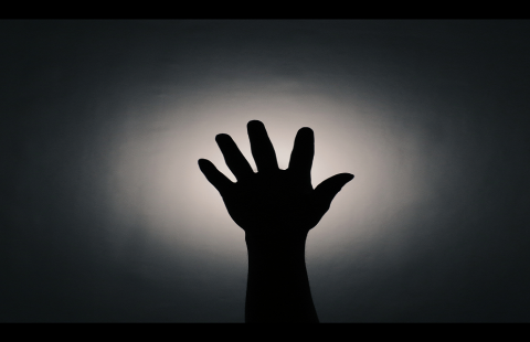 Mustalla taustalla keskellä valoisampi kohta jonka edessä ylöspäin kurottava käsi sormet harallaan.