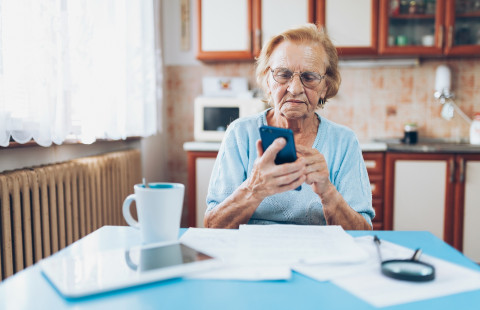 Ikäihminen selaa matkapuhelinta keittiön pöydän ääressä.