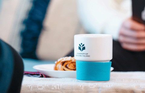 Pöydällä on kahvikuppi, jossa on yliopiston logo, sekä korvapuusti.