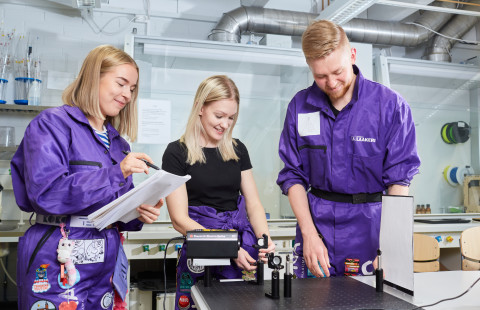 Kolme fotoniikan opiskelijaa valolaboratoriossa violetit haalarit yllään