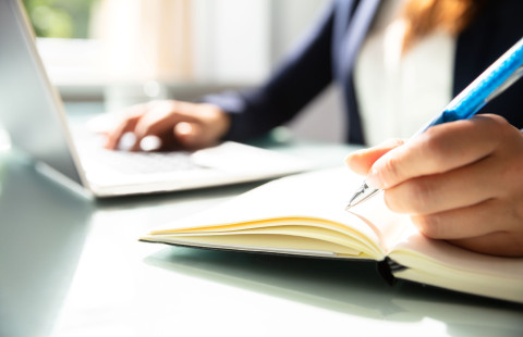 Henkilö kirjoittaa muistivihkoon taustalla kannettava tietokone, jonka näppäimistöllä toinen käsi on.