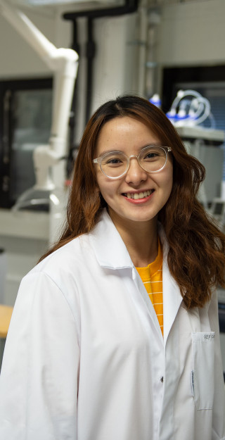 Young woman in a lab coat smiling, laboratory in the background.