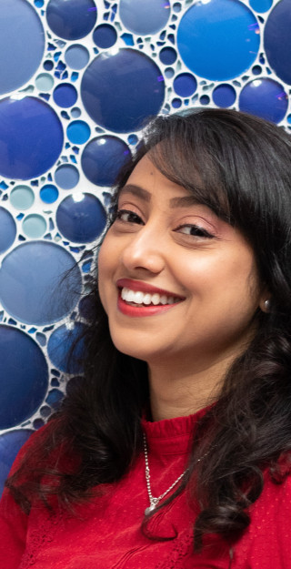 Nainen punaisessa paidassa hymyilee kameralle takanaan sinisistä lasiympyröistä muodostuva tausta.