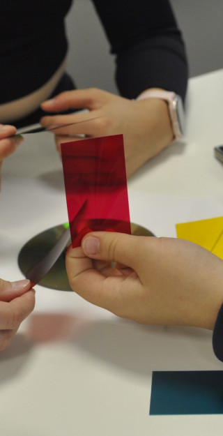 Kuvassa lasten käsiä, kun he ovat kokeilemassa erilaisia värifilttereitä tutkiessaan, kuinka valo käyttäytyy heijastuessaan CD-levyn pinnasta.